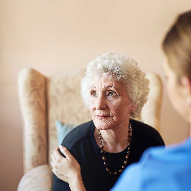Older adult arranging labeled clothing, toiletries, photos, and a small lamp in an assisted living room.